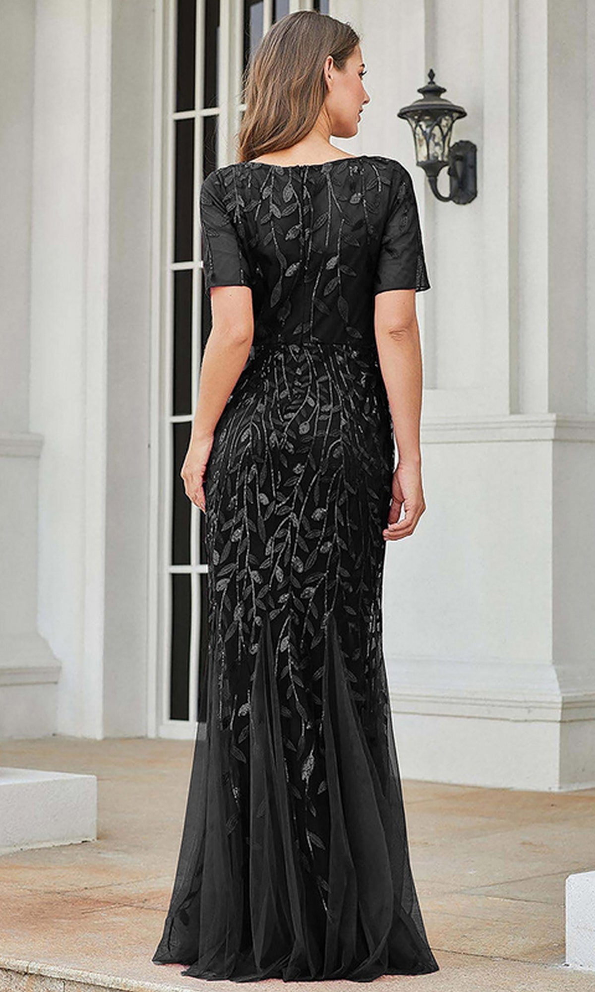 A woman stands on a porch, facing away, wearing the Short-Sleeve Modest Long Mermaid Prom Dress 7707—a black formal gown with elegant embroidery and a leaf pattern.